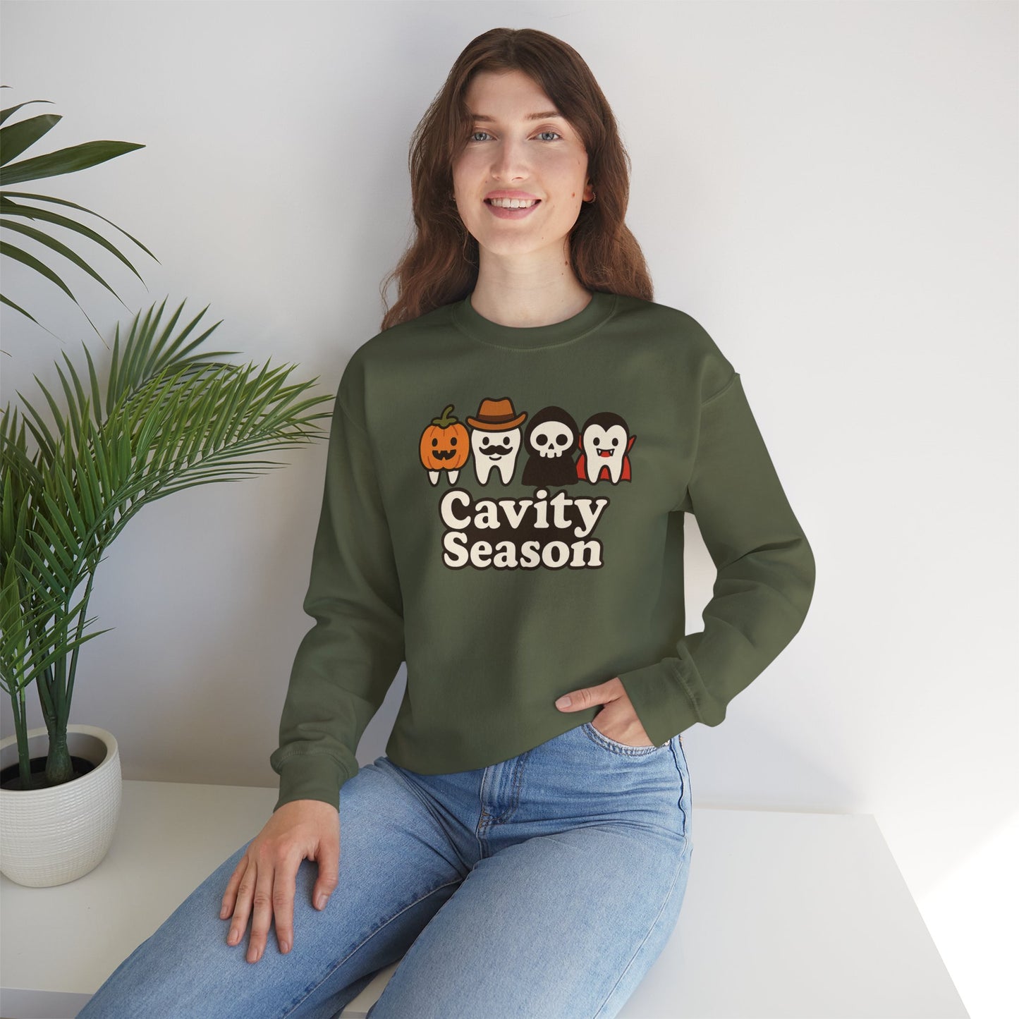 Woman wearing a dark green "Cavity Season" Halloween sweatshirt with four cartoon teeth dressed in festive costumes – pumpkin, cowboy, skeleton, and vampire – perfect for fall and spooky season celebrations.