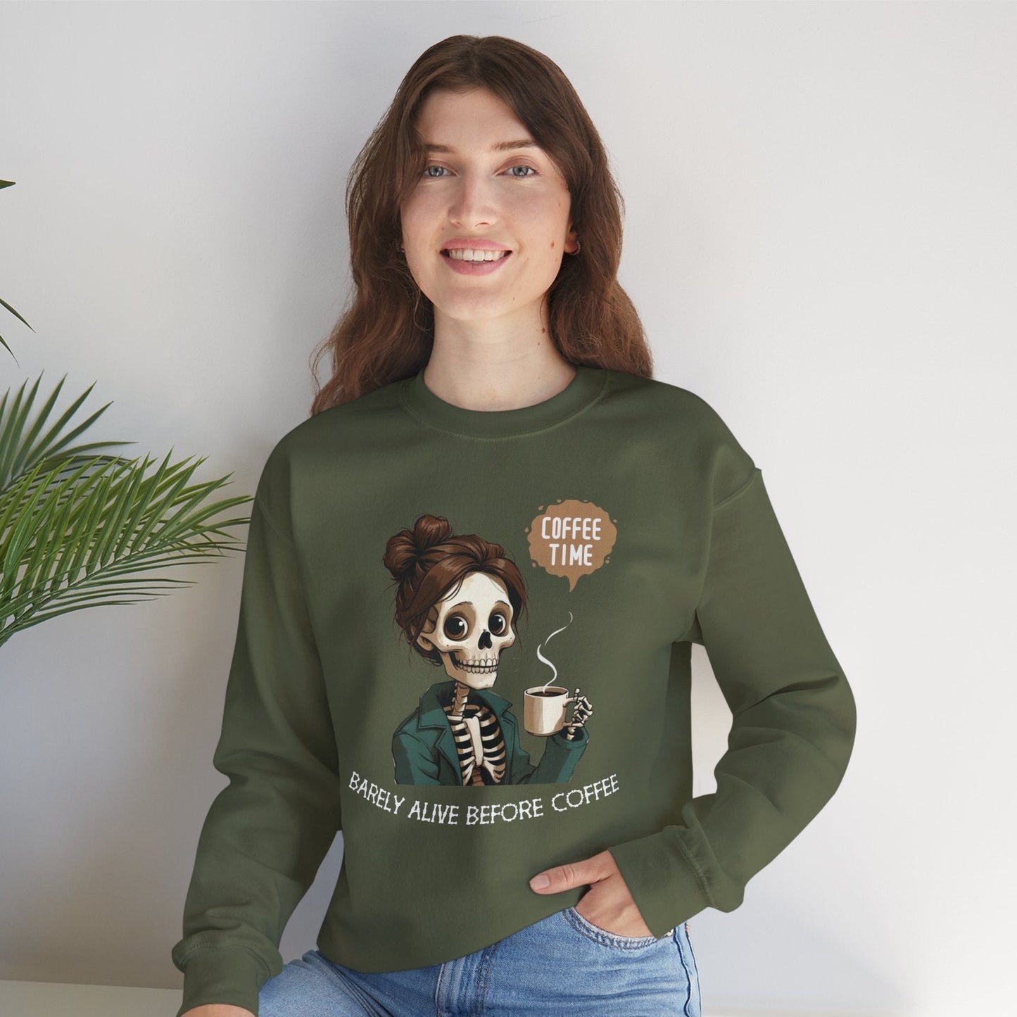 "Smiling woman wearing an olive green sweatshirt with a skeleton holding coffee and text that reads 'Barely Alive Before Coffee – Coffee Time' – funny coffee lover crewneck"