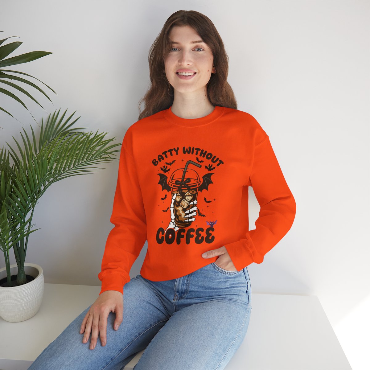 Woman wearing bright orange Halloween sweatshirt with “Batty Without Coffee” design featuring a skeleton hand holding iced coffee with bat wings; unisex crewneck made from ethically grown US cotton.
