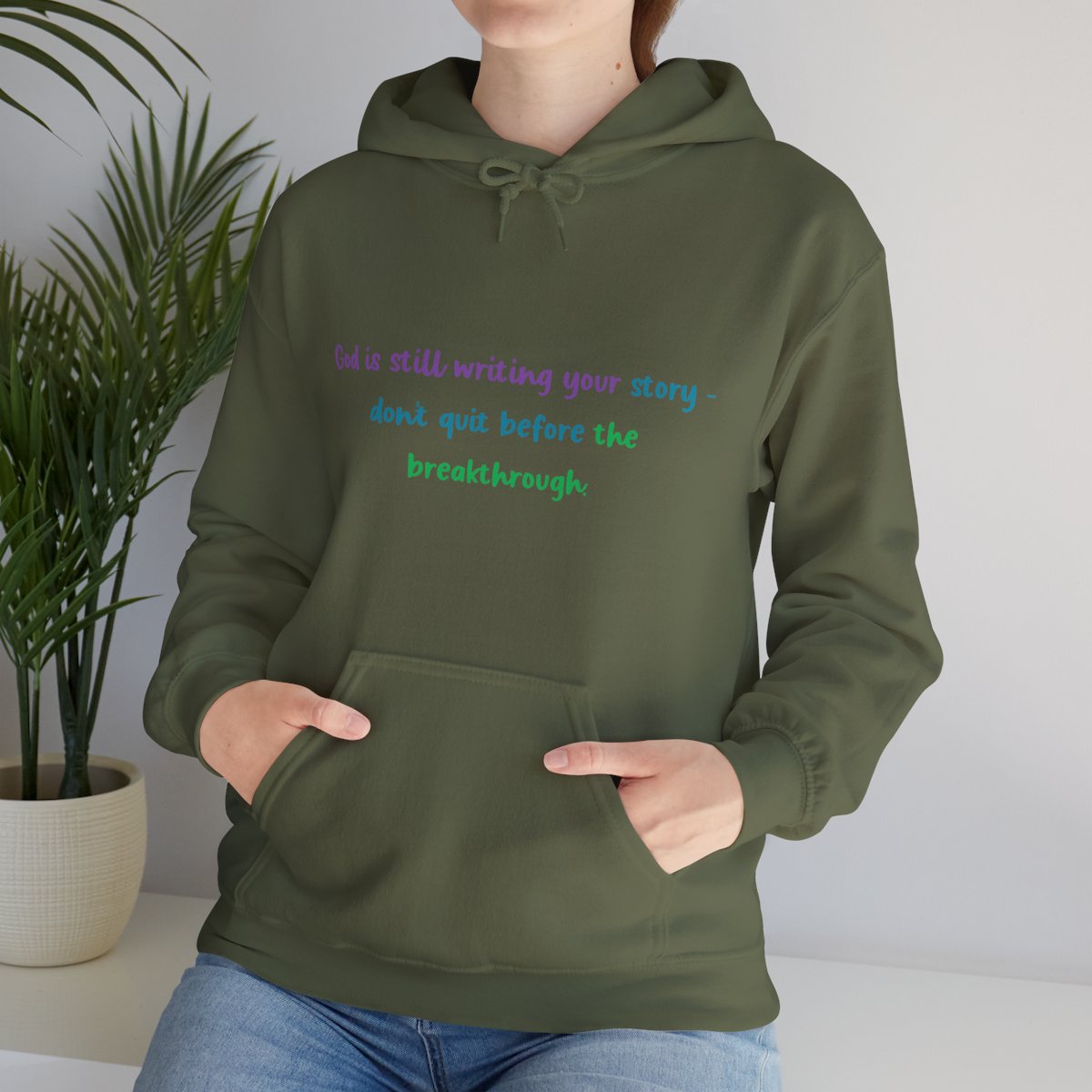 Woman wearing an olive green unisex hoodie with the inspirational quote "God is still writing your story – don't quit before the breakthrough" printed in purple, blue, and green fonts. The hoodie features a kangaroo pocket and matching drawstring hood, styled for warmth and comfort in a casual indoor setting.