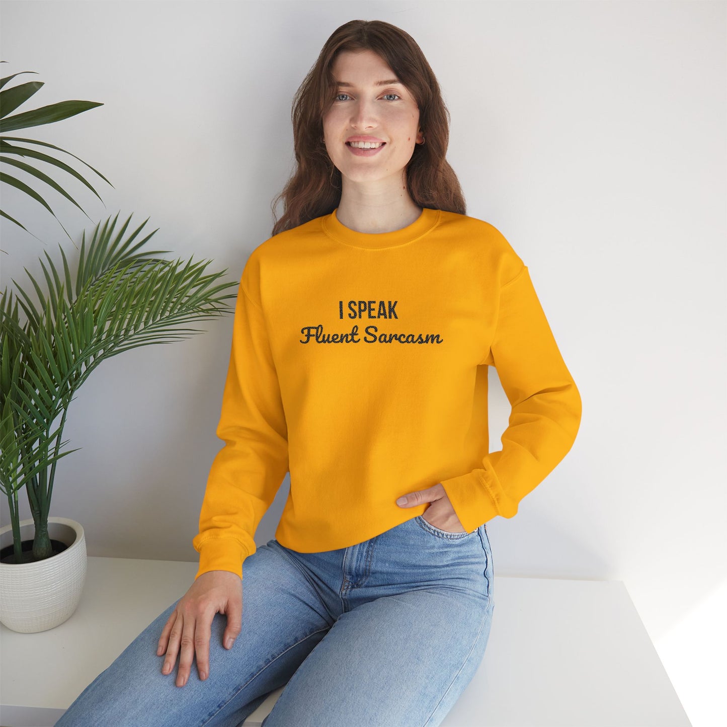 Woman wearing vibrant yellow crewneck sweatshirt with "I Speak Fluent Sarcasm" embroidered on chest in black lettering, styled with blue jeans in modern minimalist setting