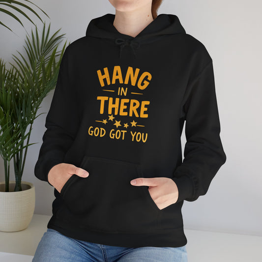 "Person wearing black Hang In There God Got You hoodie with golden inspirational text, featuring kangaroo pocket and adjustable drawstring hood, standing against white wall with decorative plant"