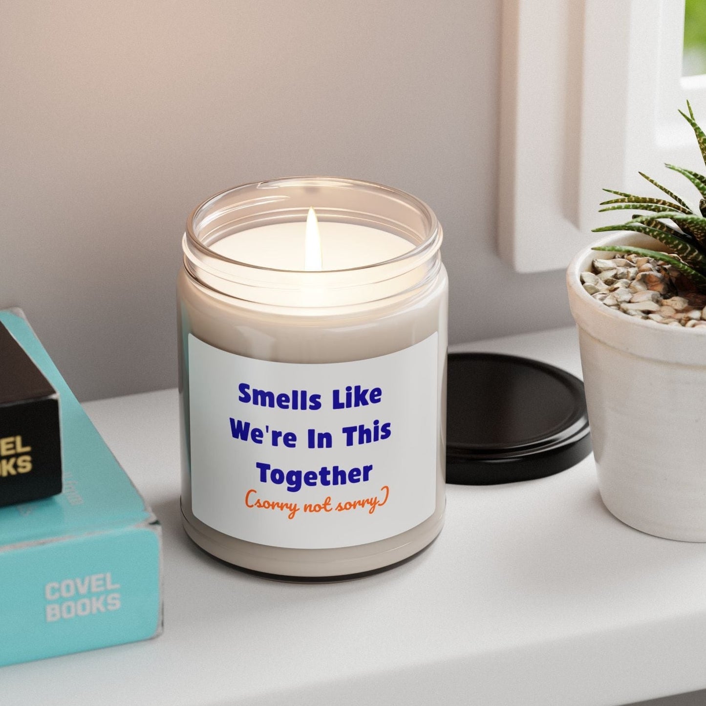 White glass jar soy candle with "Smells Like We're In This Together" blue and orange text label on desk with succulent plant and books, 9oz scented candle, funny friendship gift, home decor accent