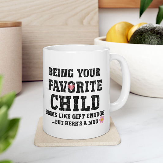 Humorous white ceramic mug with black text reading "Being Your Favorite Child Seems Like Gift Enough...But Here's A Mug" with small pink heart and flower accents