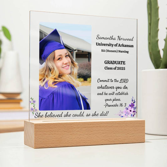 Personalized acrylic graduation plaque with the graduate's name, school, degree, graduation year, and the quote 'She believed she could, and she did,' followed by Proverbs 16:3, displayed on a wooden LED base with 7 color-changing options.
