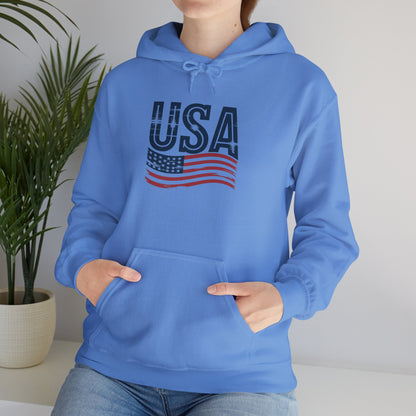 Person wearing a light blue unisex hoodie featuring a distressed “USA” text and waving American flag design, with hands in a spacious front pocket.