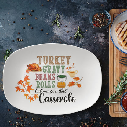 Oval white serving platter with colorful Thanksgiving design featuring text "Turkey Gravy and Beans Rolls Let Me See That Casserole" with turkey illustration, fall leaves, and food icons on dark rustic background with rosemary sprigs and peppercorns
