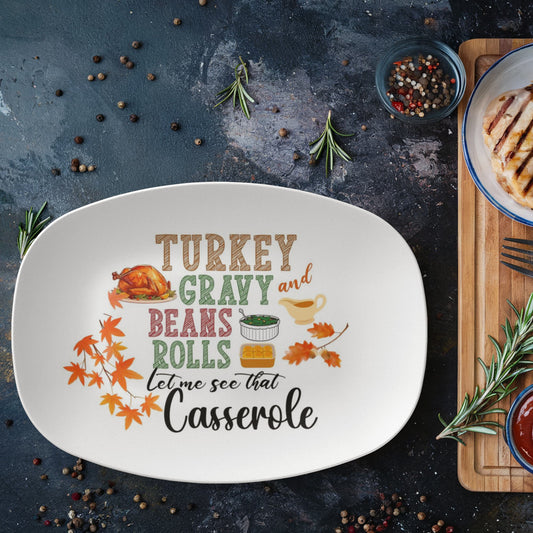Oval white serving platter with colorful Thanksgiving design featuring text "Turkey Gravy and Beans Rolls Let Me See That Casserole" with turkey illustration, fall leaves, and food icons on dark rustic background with rosemary sprigs and peppercorns