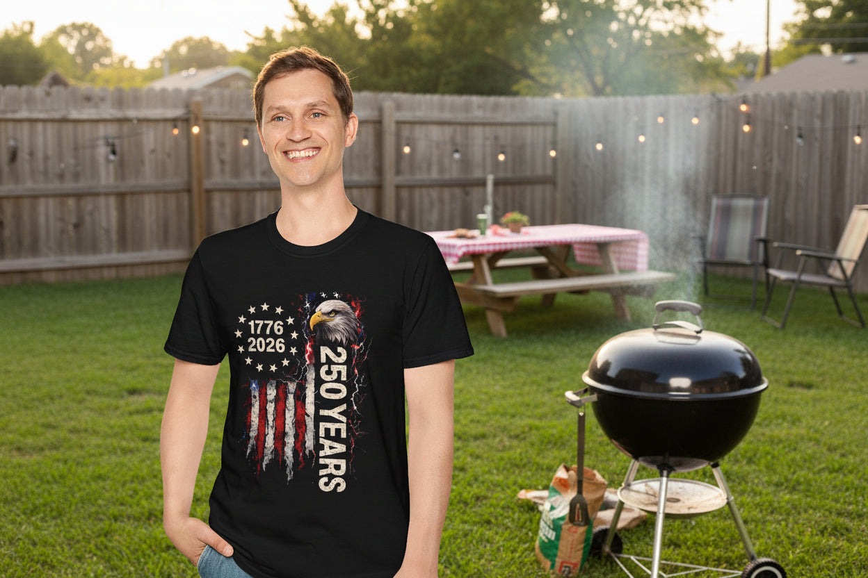 Man wearing a black Gildan Softstyle 64000 unisex t-shirt with a bald eagle and distressed American flag graphic, showing dates 1776–2026 and the text "250 Years" celebrating the USA's 250th anniversary, standing outdoors at a backyard BBQ