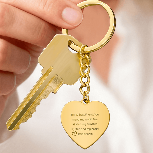 A gold heart-shaped engraved keychain attached to a gold key, held in a hand. The heart charm displays a sentimental message and features polished stainless steel with an 18K gold finish.