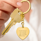 A gold heart-shaped engraved keychain attached to a gold key, held in a hand. The heart charm displays a sentimental message and features polished stainless steel with an 18K gold finish.