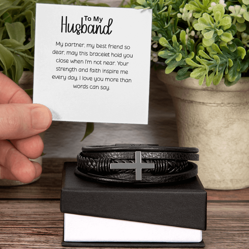 Black braided leather men's bracelet with stainless steel cross displayed on black gift box with "To My Husband" romantic message card held by hand against rustic wooden background