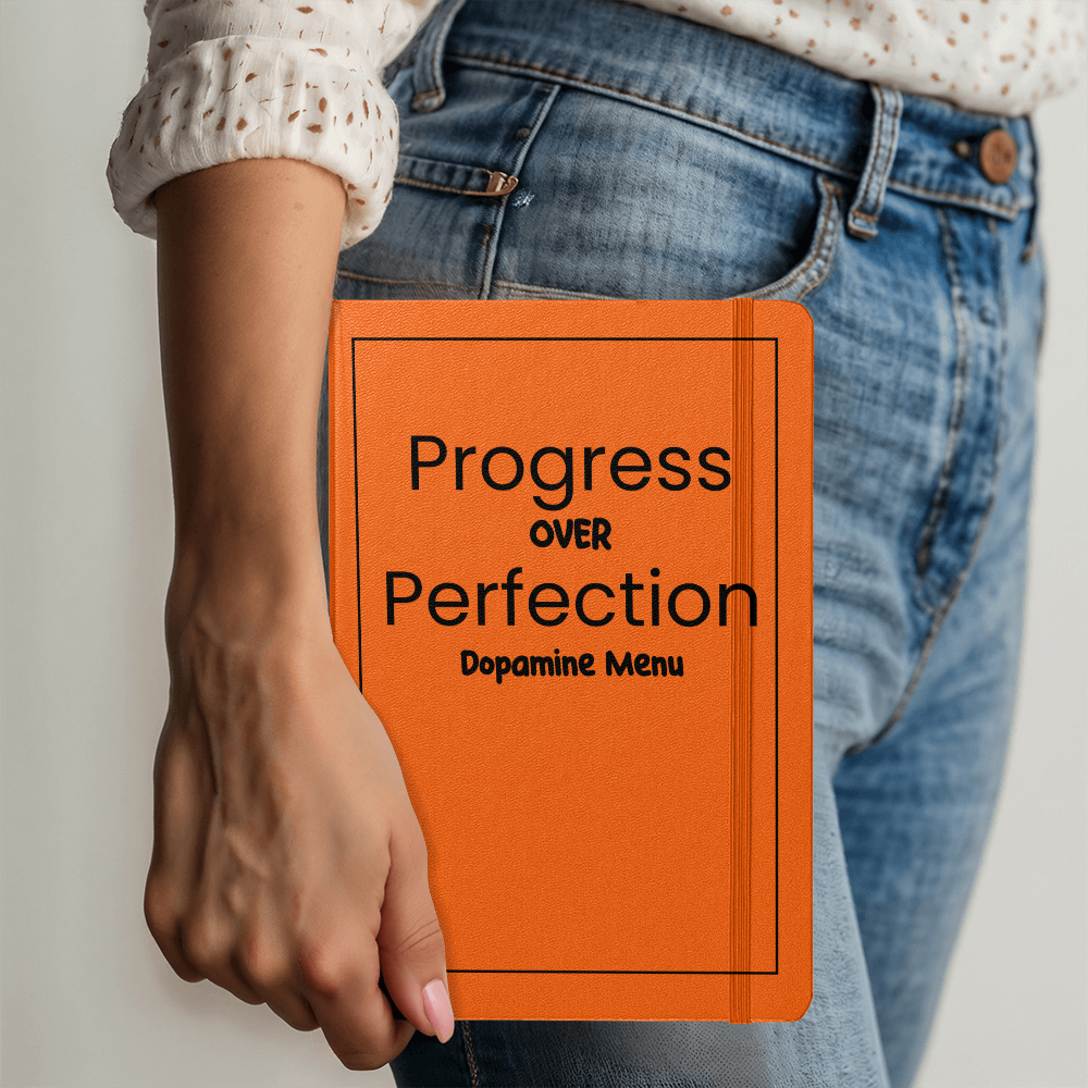 Orange hardcover journal with "Progress Over Perfection Dopamine Menu" text held against denim jeans, featuring elastic closure and ribbon marker for goal tracking and daily planning