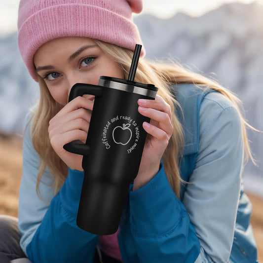 Teacher holding black stainless steel tumbler with the words “Caffeinated and Ready to Inspire Minds” laser engraved on the front, featuring matte finish, handle, lid, and straw.