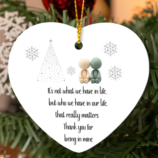Heart-shaped high gloss MDF ornament with two simple character figures sitting together, snowflakes, a dotted Christmas tree, and the quote “It’s not what we have in life but who we have that really matters. Thank you for being in mine,” hanging on a Christmas tree with a gold cord.
