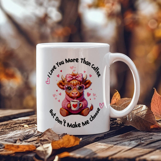 White ceramic coffee mug featuring cute highland cow wearing pink bow and glasses, holding heart mug, with text "I Love You More Than Coffee But Don't Make Me Choose" surrounded by pink hearts