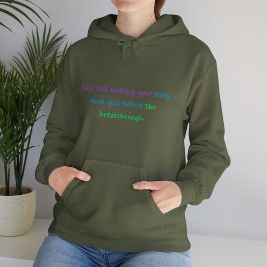 Woman wearing an olive green unisex hoodie with the inspirational quote "God is still writing your story – don't quit before the breakthrough" printed in purple, blue, and green fonts. The hoodie features a kangaroo pocket and matching drawstring hood, styled for warmth and comfort in a casual indoor setting.